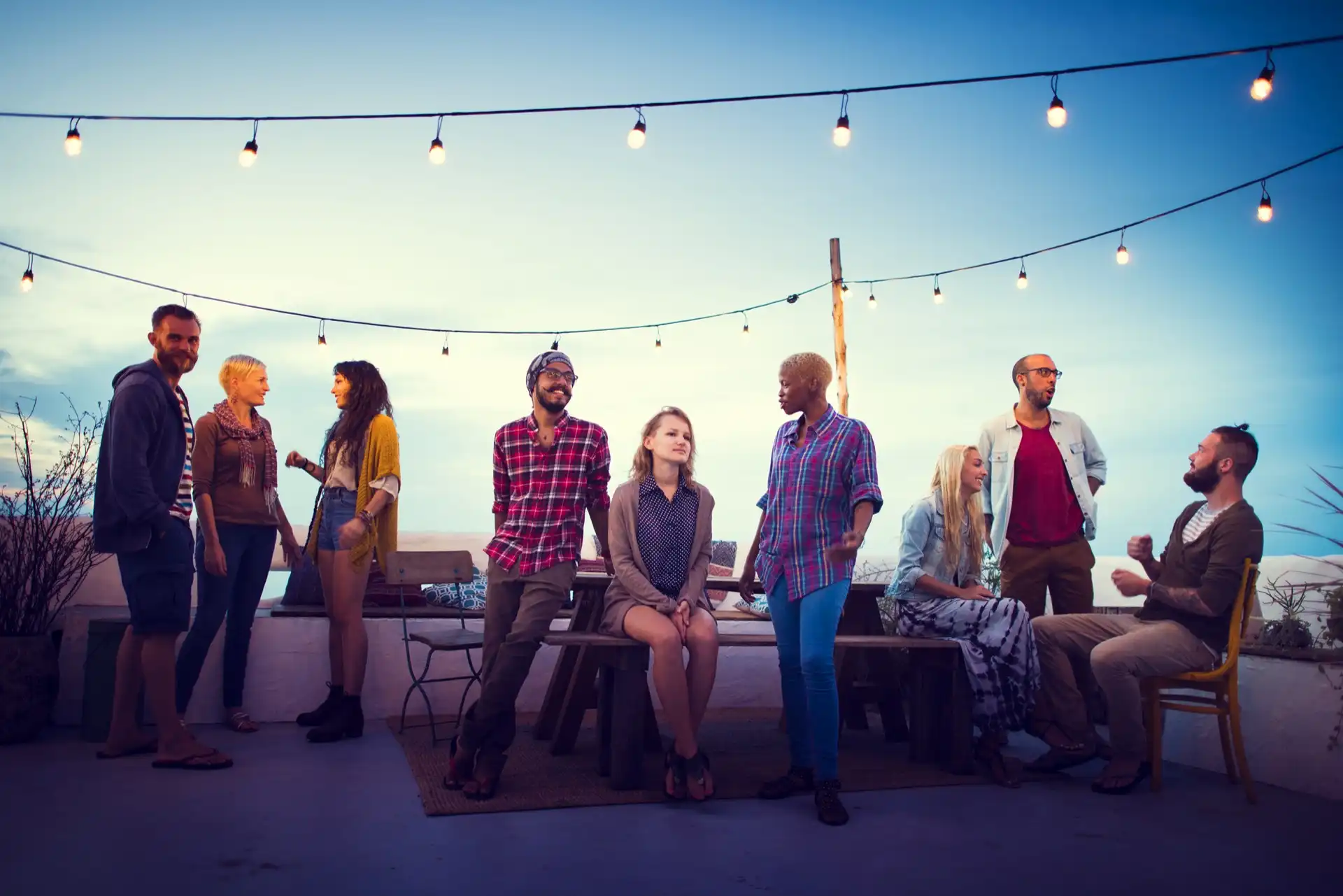 Young adults of various ethnicities socializing on a rooftop patio under rope lights at dusk.